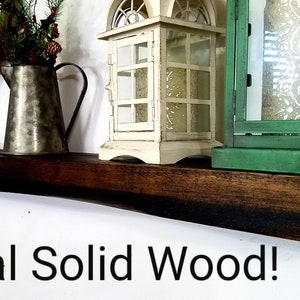 May include: A dark brown wooden shelf with a rustic finish. The shelf is decorated with a metal pitcher, a white lantern, a green lantern, and a wooden sign that says "Bless this Nest".
