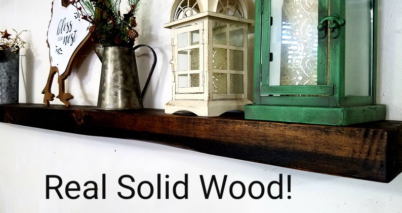 May include: A dark brown wooden shelf with a rustic finish. The shelf is decorated with a metal pitcher, a white lantern, a green lantern, and a wooden sign that says "Bless this Nest".