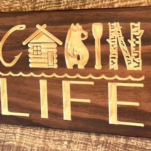 May include: Wooden sign with the words "CABIN LIFE" carved into it. The design includes a cabin, bear, paddle, and birch tree. The sign is made of dark brown wood with lighter wood used for the carved elements.