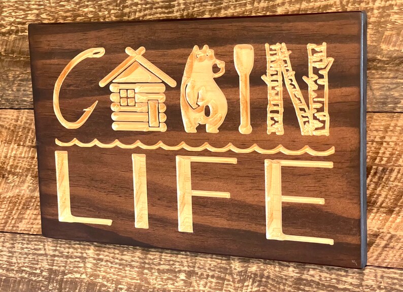 May include: Wooden sign with the words "CABIN LIFE" carved into it. The design includes a cabin, bear, paddle, and birch tree. The sign is made of dark brown wood with lighter wood used for the carved elements.