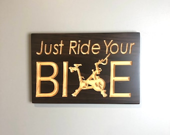 May include: A dark brown wooden sign with the text "Just Ride Your Bike" in a light yellow color. A stylized image of a stationary bike is carved into the wood.
