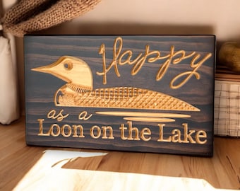 Può includere: Un cartello di legno con il testo "Happy as a Loon on the Lake" inciso su di esso. Il cartello è marrone scuro con un loon e un testo dorati.