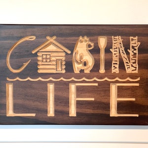 May include: Wooden sign with the words "CABIN LIFE" carved into the surface. The design includes a fishing hook, cabin, bear, paddle, and trees. The sign is made of dark brown wood with lighter wood used for the carved elements.