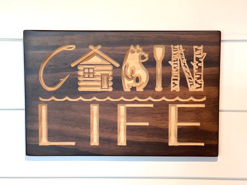 May include: Wooden sign with the words "CABIN LIFE" carved into the surface. The design includes a fishing hook, cabin, bear, paddle, and trees. The sign is made of dark brown wood with lighter wood used for the carved elements.