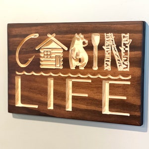 May include: A wooden sign with the words "CABIN LIFE" carved into it. The letters and accompanying images are light-colored against a dark brown background. The images include a fishing hook, a cabin, a bear, a paddle, and trees.