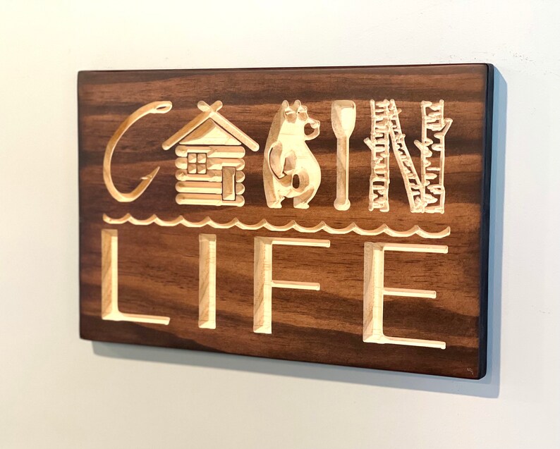 May include: A wooden sign with the words "CABIN LIFE" carved into it. The letters and accompanying images are light-colored against a dark brown background. The images include a fishing hook, a cabin, a bear, a paddle, and trees.