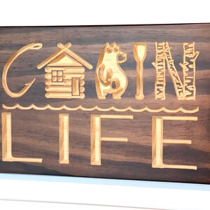 May include: Wooden sign with the words "CABIN LIFE" carved into the surface. The letters are accompanied by carved images of a cabin, bear, paddle, and trees. The sign is stained a dark brown color and mounted on a white wall.