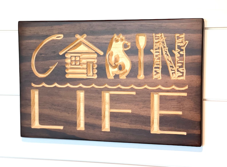 May include: Wooden sign with the words "CABIN LIFE" carved into the surface. The letters are accompanied by carved images of a cabin, bear, paddle, and trees. The sign is stained a dark brown color and mounted on a white wall.