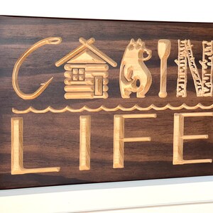 May include: Wooden sign with the words "CABIN LIFE" carved into it. The sign features a fishing hook, a cabin, a bear, a paddle, and trees. The sign is made of dark brown wood with lighter wood used for the carved elements.