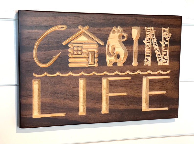May include: Wooden sign with the words "CABIN LIFE" carved into it. The sign features a fishing hook, a cabin, a bear, a paddle, and trees. The sign is made of dark brown wood with lighter wood used for the carved elements.