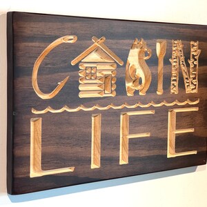 May include: Wooden sign with the words "CABIN LIFE" carved into the wood. The letters and images are light-colored against a dark brown background. The design includes a cabin, bear, spoon, and trees.