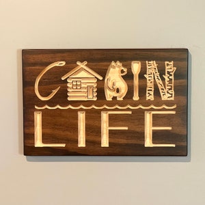 May include: Wooden sign with the words "Cabin Life" carved into a dark brown background. The design includes a fishing hook, cabin, bear, paddle, birch trees, and waves. A decorative piece for a cabin or lake house.
