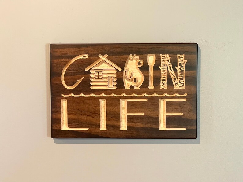 May include: Wooden sign with the words "Cabin Life" carved into a dark brown background. The design includes a fishing hook, cabin, bear, paddle, birch trees, and waves. A decorative piece for a cabin or lake house.