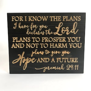May include: Black wooden sign with gold lettering that reads "For I know the plans I have for you declares the Lord plans to prosper you and not to harm you plans to give you hope and a future Jeremiah 29:11".