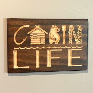 May include: Wooden sign with the words "CABIN LIFE" carved into the surface. The letters are accompanied by carved images of a cabin, bear, spoon, and trees. The sign is rectangular and made of dark brown wood.