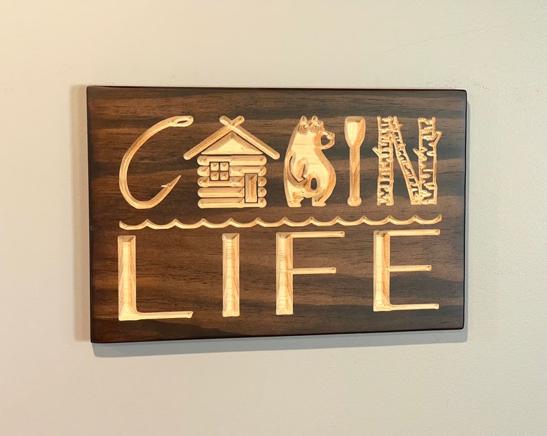 May include: Wooden sign with the words "CABIN LIFE" carved into the surface. The letters are accompanied by carved images of a cabin, bear, spoon, and trees. The sign is rectangular and made of dark brown wood.