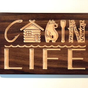 May include: Wooden sign with the words "CABIN LIFE" carved into a dark brown background. The design includes a fishing hook, cabin, bear, paddle, ladder, and waves. The letters and images are light brown.