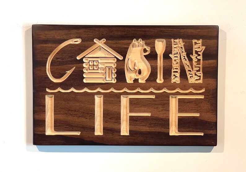 May include: Wooden sign with the words "CABIN LIFE" carved into a dark brown background. The design includes a fishing hook, cabin, bear, paddle, ladder, and waves. The letters and images are light brown.