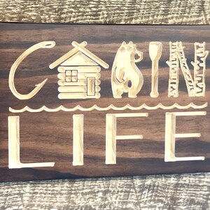 May include: A wooden sign with the words "CABIN LIFE" carved into it. The sign features a cabin, bear, paddle, ladder, and trees. The sign is made of dark brown wood with light brown carved details.