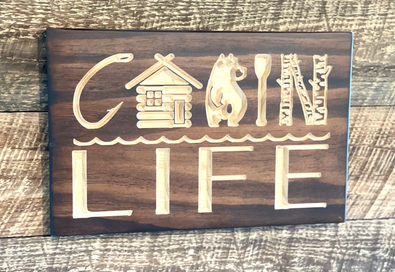 May include: A wooden sign with the words "CABIN LIFE" carved into it. The sign features a cabin, bear, paddle, ladder, and trees. The sign is made of dark brown wood with light brown carved details.