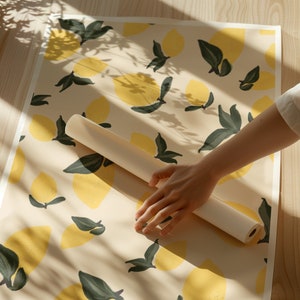 Könnte beinhalten: Eine weiße Rolle Geschenkpapier mit einem Zitronen-Druckmuster. Das Papier wird auf einer hellen Holzoberfläche abgewickelt.
