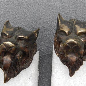 May include: Two antique bronze-toned lion head sculptures. Each sculpture features detailed facial features, including a prominent nose, open mouth, and pointed ears. The lion heads are mounted on white foam.