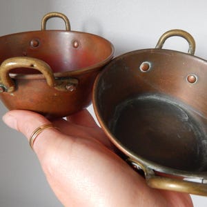 Op de afbeelding: Twee kleine koperen pannen met messing handvatten. De pannen hebben een afgeronde vorm en zijn geklonken. De handvatten zijn gebogen. Het koper heeft een warme, roodbruine tint, wat duidt op leeftijd en gebruik. Deze pannen zijn waarschijnlijk voor decoratieve doeleinden of kleinschalig koken.