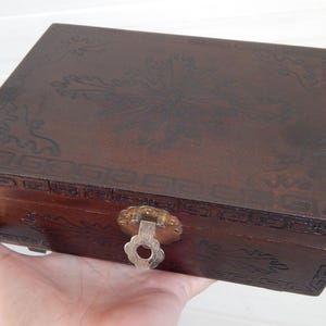 May include: A rectangular, dark brown wooden box with a decorative floral design carved into the lid. The box has a metal clasp and is held in a hand. The box is likely used for storage.