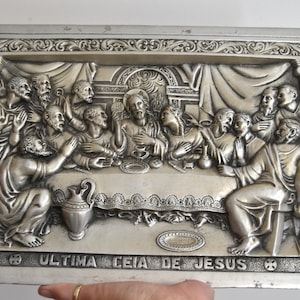 May include: A rectangular silver-toned metal wall plaque featuring a high-relief depiction of The Last Supper. The scene shows figures seated at a long table with architectural columns on the sides. The text ULTIMA CEIA DE JESUS is embossed at the bottom.