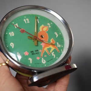 Puede incluir: Reloj despertador vintage con esfera verde, ilustración de ciervo de dibujos animados y la palabra "HERO". El reloj tiene un marco de metal plateado y una base azul. Las manecillas son doradas y rojas.