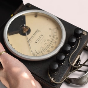 Puede incluir: Un multímetro vintage negro y plateado con un dial redondo etiquetado como "VOLTS" y múltiples botones. El multímetro tiene un cable verde y rojo conectado a él.