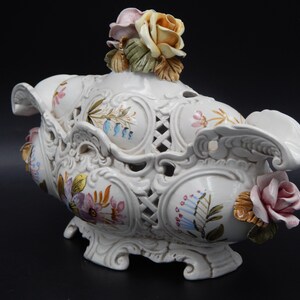 Puede incluir: Un cuenco decorativo de cerámica blanca con tapa, con intrincados diseños calados y florales. El cuenco está adornado con flores pintadas a mano en tonos rosa, amarillo y azul. La tapa está rematada con un grupo de rosas de cerámica.