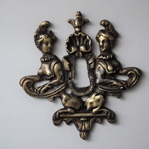 May include: A gold-toned metal keyhole cover with two female figures flanking a keyhole. The figures are wearing elaborate headdresses and are holding scrolls.