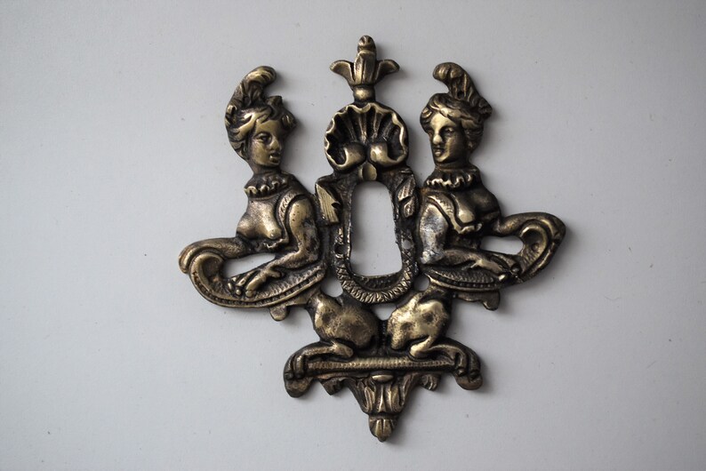 May include: A gold-toned metal keyhole cover with two female figures flanking a keyhole. The figures are wearing elaborate headdresses and are holding scrolls.