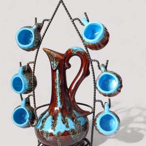 Op de afbeelding: Een vintage keramische kan en kopjes set. De kan is bruin met blauwe accenten en een gebogen handvat. Zes kleine kopjes met blauwe binnenkant hangen aan een smeedijzeren standaard. Glanzende afwerking.