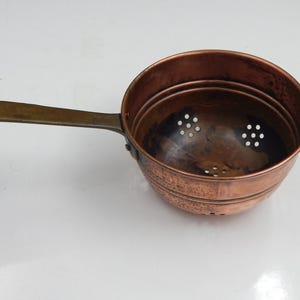 Può includere: Una scolapasta in rame con un lungo manico in ottone. La scolapasta ha una ciotola rotonda con più fori di drenaggio. Il manico ha un foro per appenderla. Il rame ha un colore marrone-rossastro caldo.