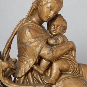 May include: A gold-colored statue depicting a mother and child. The mother figure is draped in flowing robes and cradles the child. The statue has an aged appearance with visible wear and tear, adding to its antique aesthetic.