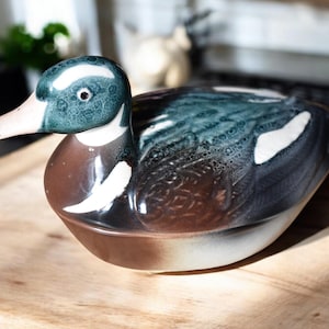 May include: A ceramic duck figurine with a brown, white, and green glaze. The duck has a white belly and a green head with a white stripe.