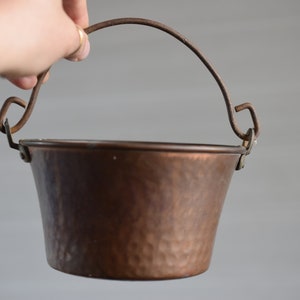 Può includere: Un piccolo secchiello di rame con finitura martellata e manico in filo metallico. Il secchiello è vuoto e ha un aspetto rustico.