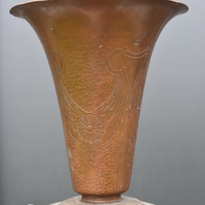 May include: A hammered copper vase with an ornate design. The vase has a wide mouth and a narrow base. It is decorated with a floral pattern and a geometric design.