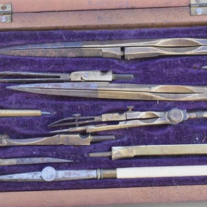 Può includere: Una collezione di antichi strumenti da disegno, tra cui compassi, divisori e un goniometro, disposti in una scatola di legno con rivestimento in velluto viola. Gli strumenti sono realizzati in ottone e acciaio, mostrando segni di età e utilizzo.