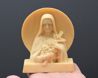 May include: A white figurine of a woman holding a bouquet of flowers and a cross. The figurine is on a white base with a rounded back.