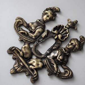 May include: A gold-toned metal decorative switch plate featuring two stylized figures with ornate details. The figures are positioned around a central opening for a light switch.
