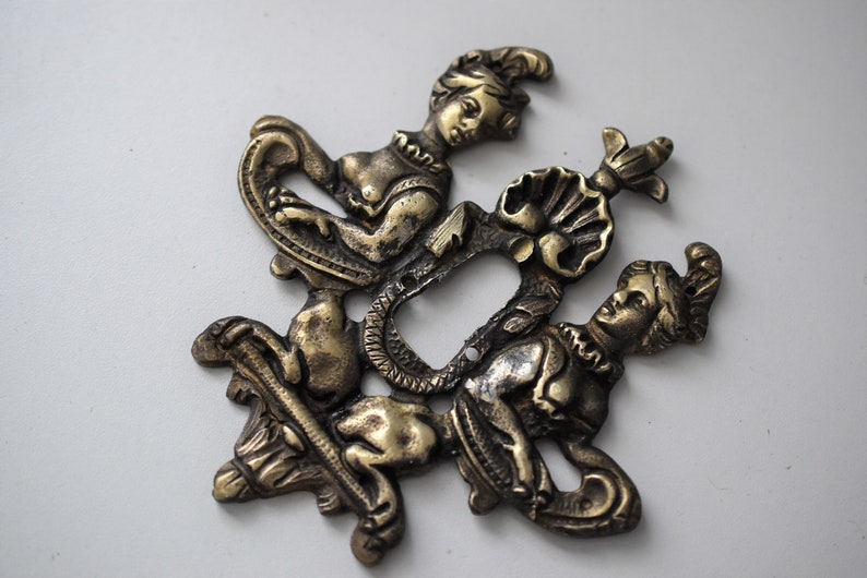 May include: A gold-toned metal decorative switch plate featuring two stylized figures with ornate details. The figures are positioned around a central opening for a light switch.