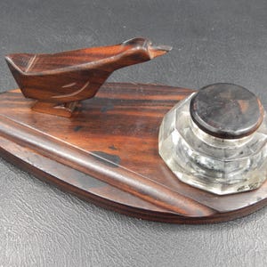 Pode incluir: Um conjunto de tinteiro de madeira vintage com uma figura de pássaro esculpida e um tinteiro de vidro facetado com tampa escura. O conjunto é feito de madeira marrom escura e fica em uma base oval.