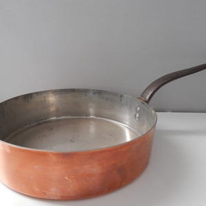 May include: A copper-colored frying pan with a long, dark handle. The pan has a wide, shallow bowl and a shiny interior. The handle is curved and appears to be made of a dark metal. The pan is likely used for cooking.