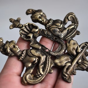 May include: A gold-toned metal ornament featuring three figures in a stylized design. The figures are depicted in a dynamic pose, with one figure riding a mythical creature. The ornament has a detailed and intricate design.