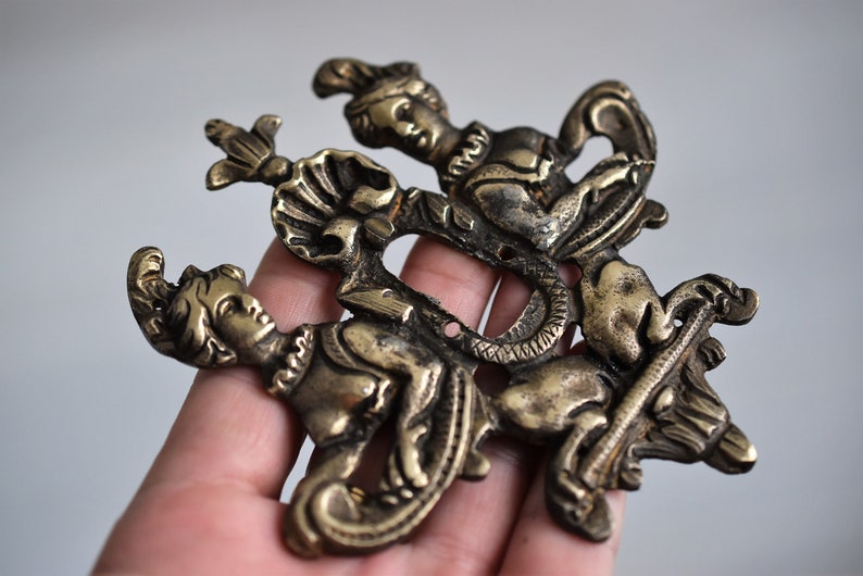 May include: A gold-toned metal ornament featuring three figures in a stylized design. The figures are depicted in a dynamic pose, with one figure riding a mythical creature. The ornament has a detailed and intricate design.