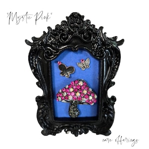 May include: A black framed display with a blue background featuring a mushroom and two butterfly brooches. The mushroom has a pink and clear crystal top. The butterflies are black and silver. The text "Mystic Pink" and "rare offerings" are in the image.