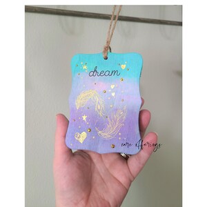 May include: A wooden ornament with a teal and purple ombre background. The ornament features gold glitter, stars, hearts, and two feathers. The word "dream" is written in black on the ornament. The bottom of the ornament says "rare offerings".